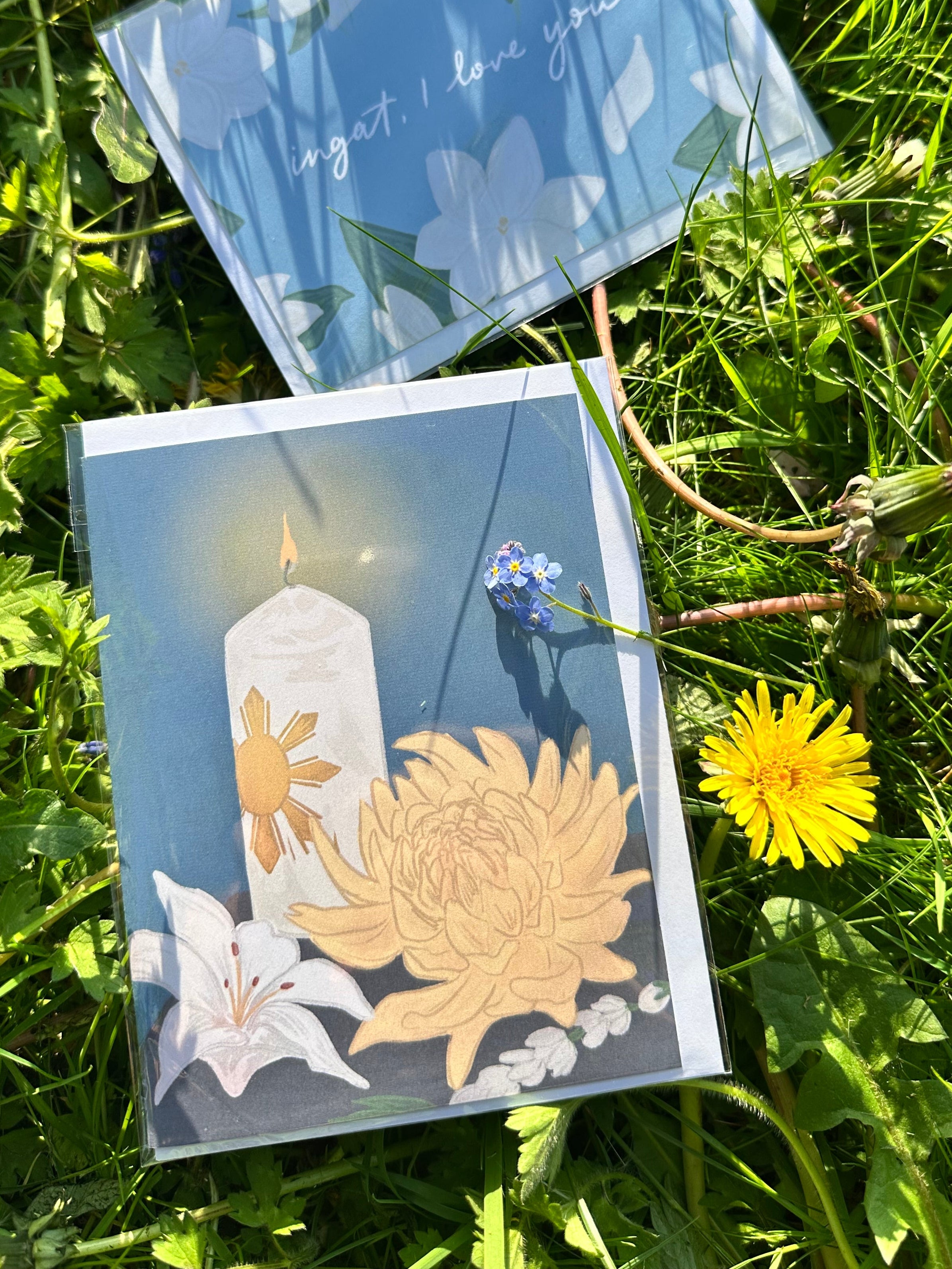 Pakikidalamhati card laying on the grass. Dandelions and a sprig of forget-me-not can be seen in frame.